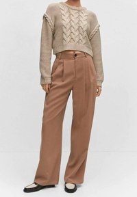 Beiger Strickpullover mit strukturiertem Zopfmuster, kombiniert mit hochtaillierter, weiter brauner Hose. Schwarze und cremefarbene Schuhe vervollständigen das Outfit.