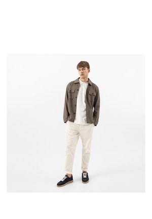 Jeune homme debout les mains dans les poches, portant une veste marron, un t-shirt blanc, un pantalon beige et des baskets noires sur fond blanc.