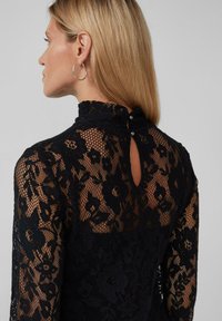 Robe en dentelle noire avec un col haut, un motif floral et un détail de boutons à l'arrière. Texture transparente et manches longues.