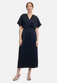 Navyblaues Maxikleid mit einem Wickelstil-Front, schwingenden Ärmeln und einem seitlichen Schlitz; kombiniert mit silbernen Sandalen mit Absatz und Riemchen.
