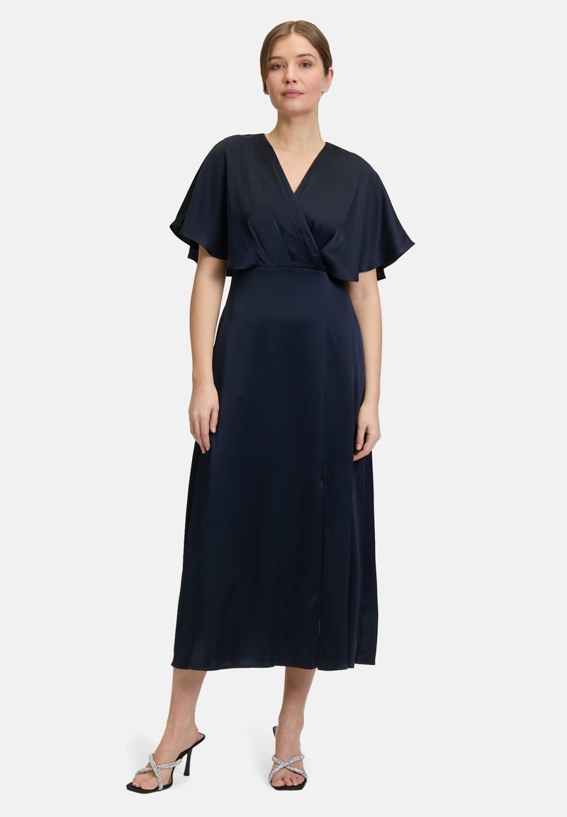 Navyblaues Maxikleid mit einem Wickelstil-Front, schwingenden Ärmeln und einem seitlichen Schlitz; kombiniert mit silbernen Sandalen mit Absatz und Riemchen.