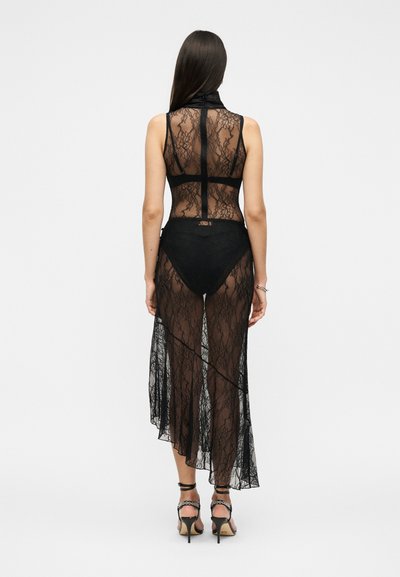 Femme debout, portant une robe noire en dentelle transparente sans manches avec un col montant, associée à des sous-vêtements noirs et des sandales à talons hauts.