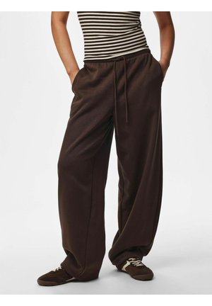 Personne portant un pantalon de survêtement large marron foncé, les mains dans les poches, et des baskets marron, associée à un haut rayé noir et blanc.
