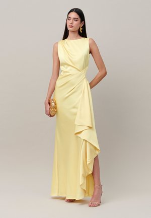 Mujer con cabello largo y oscuro que lleva un vestido de satén amarillo pálido sin mangas con detalle drapeado, sosteniendo un bolso de mano dorado y llevando pendientes y tacones dorados.