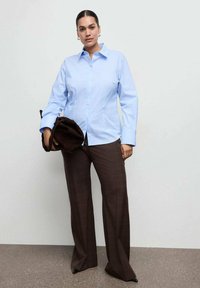 Svetlomodrá košeľa s dlhým rukávom a golierom, doplnená hnedými nohavicami s širokými nohavicami v kockovanom vzore. Pod pazuchou tmavohnedá taška.