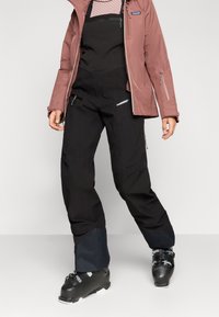 Melni ūdensizturīgi slēpošanas bikses ar sašaurinātu piegriezumu, sānu rāvējslēdzējiem un kontrastējošu zilu akcentu pie apakšmalām, apvienotas ar melniem slēpošanas zābakiem.