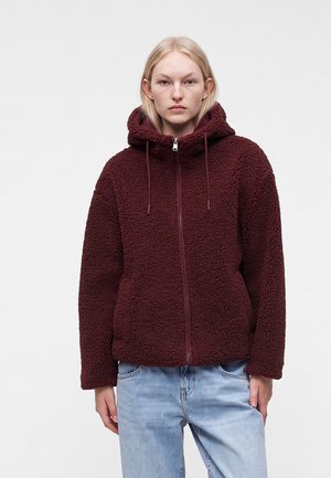 Femme portant un sweat à capuche zippé en polaire bordeaux avec une capuche et un jean bleu clair, debout devant un fond uni.