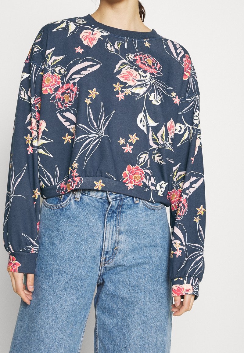 Personne portant un haut à manches longues bleu marine à motifs floraux avec des fleurs roses, rouges et blanches, associé à un jean taille haute bleu clair.