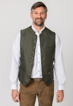 Lächelnder Mann, der traditionelle braune Wildlederhosen, weißes Hemd mit Knöpfen und dunkelgrüne gemusterte Weste mit Metallknöpfen trägt.