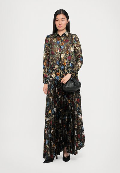 Transparante zwarte bloemenblouse met kraag, gecombineerd met een geplooide zwarte rok met veelkleurige bloemen. Houdt een zwarte clutch in de hand.
