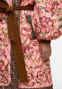 Pletený růžový kabát s květinovými vzory, hnědý kordový pásek, kontrastní lemování a knoflíky; textura látky s propracovanými vzory a akcenty.