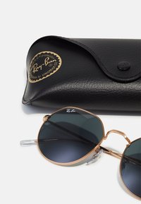 Ray-Ban aurinkolasit tummilla linsseillä, kultaisella metallikehyksellä ja läpinäkyvillä temppeleillä, mukana musta nahkakotelo, jossa on kohokuvioitu logo.