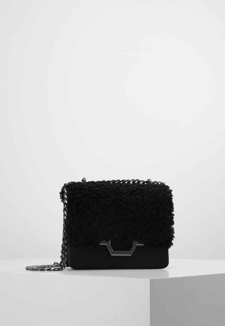 Zwarte handtas met een gestructureerd oppervlak, een zilveren kettingriem en een geometrische zilveren sluiting, tentoongesteld op een wit voetstuk tegen een grijze achtergrond.