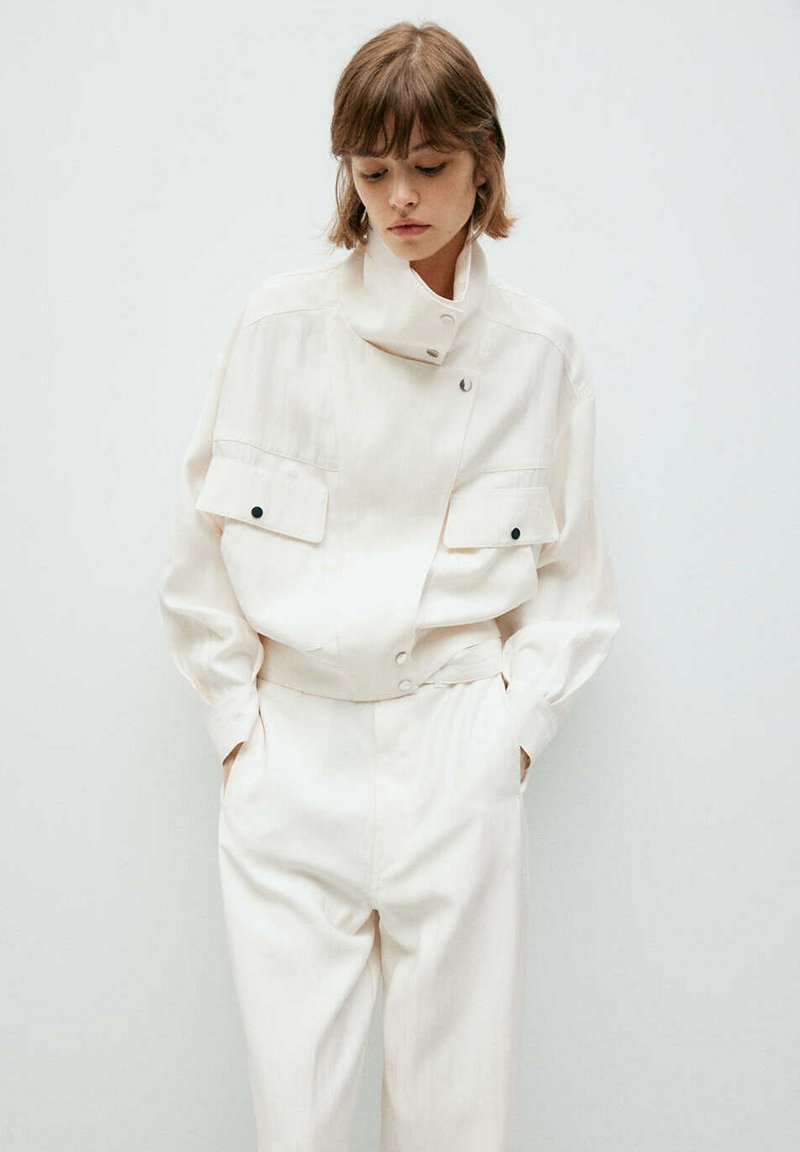 Femme aux cheveux courts portant une veste blanche à col montant avec poches avant et un pantalon blanc assorti, mains dans les poches, regardant vers le bas.