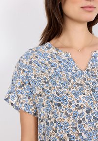 Blusa floral en crema con patrones de flores azules y grises, de mangas cortas y escote en V. Tejido suave con detalles delicados.