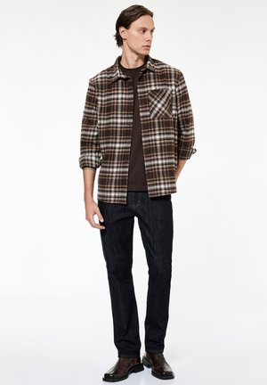 Joven con camisa marrón y blanca de cuadros sobre camiseta oscura, vaqueros oscuros y zapatos de cuero marrones, de pie frente a un fondo blanco.