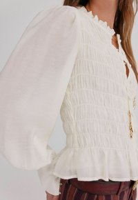 Biała bluzka z puff-sleeve, o marszczonym wykończeniu, z falbaną przy szyi i zapinana na guziki. Posiada warstwowy dół oraz naszyjnik z złotym charms w kształcie pióra.