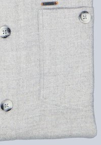 Hellgrauer Stoff mit glatter Textur, zwei silbernen Knöpfen, einer kleinen aufgesetzten Tasche und einem metallenen Logo-Akzent.