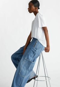 T-shirt bianco in cotone con maniche corte, abbinato a pantaloni in denim blu a vestibilità ampia con tasca con zip e una grande tasca laterale.