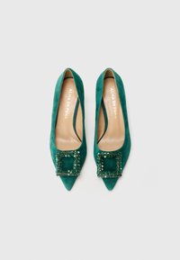 Un paio di ballerine verde scamosciato con punta appuntita e fibbie quadrate decorate da gemme, viste dall'alto su uno sfondo bianco.