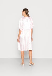 Vestido de cetim rosa com mangas bufantes, silhueta em A, padrão floral texturizado, detalhe de abertura nas costas, usado com sandálias de salto quadrado bege.