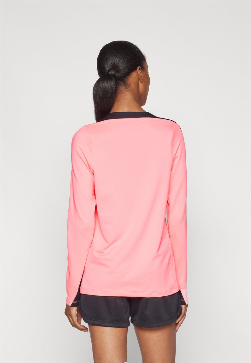 Chemise à manches longues en tissu rose vif avec un col noir et des accents latéraux, texture lisse et coupe athlétique. Portée avec un short noir.