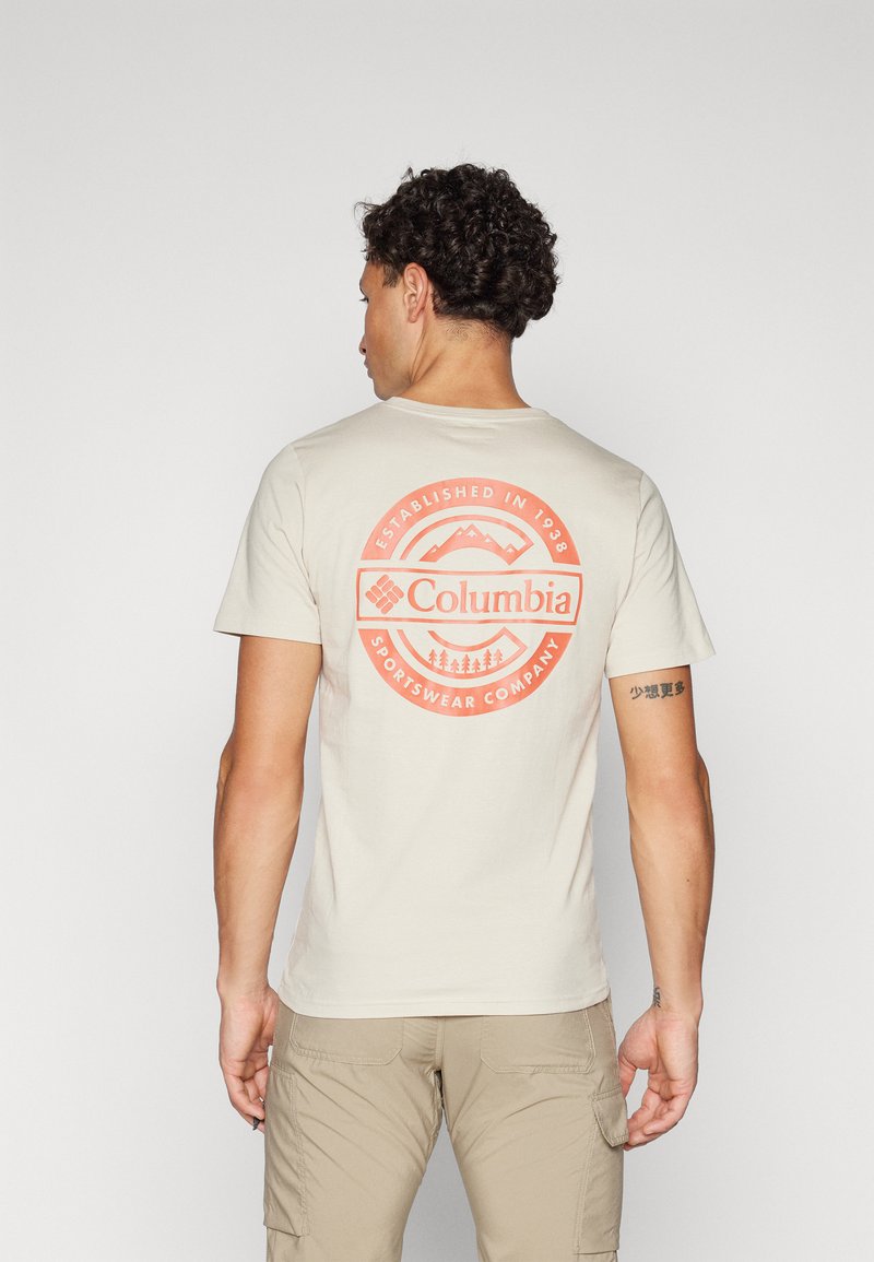 Béžové tričko s krátkym rukávom s okrúhlym červeným grafickým motívom s horami a textom "Columbia Sportswear Company" na chrbte.