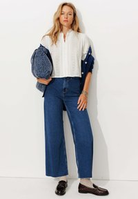 Blouse blanche texturée avec un col haut, pantalon wide-leg en jean bleu, et mocassins marron. Sac à motifs bleus porté sur un bras.