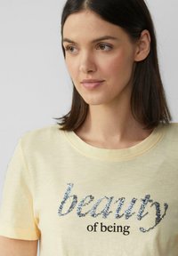 T-shirt en coton jaune clair avec un col rond et des manches courtes, arborant un texte argenté brillant "la beauté d'être" en évidence.