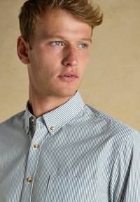 Pasiasta koszula z guzikami w jasnoniebieskim i białym kolorze, z kołnierzykiem, kieszenią na piersi i guzikami przypominającymi perły, wykonana z gładkiego materiału.