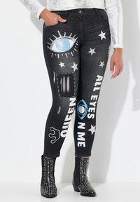 Jeans de mezclilla negra con estampados gráficos, incluyendo un ojo, estrellas y texto. Presenta un ajuste slim y un diseño de largo hasta el tobillo. Llevados con botas negras con tachuelas.