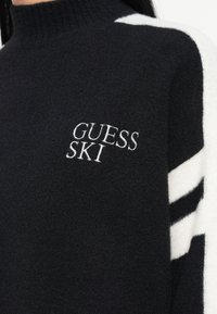 Črna flis turtleneck pulover s belimi pletenimi črtami in belim logotipom "GUESS SKI" na prsih; mehak material, prilegajoča zasnova.