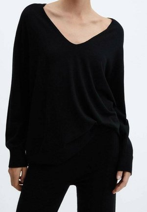 Personne portant un pull noir ample à col en V avec manches longues et un pantalon noir ajusté sur un fond clair uni.