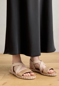 Light beige sandals featuring cross-over elastic straps and a woven upper, set against a black maxi dress on a wooden floor.