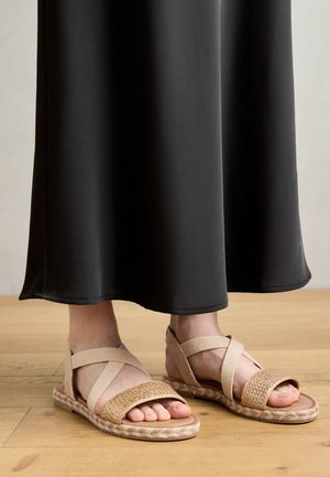 Sandales beige clair avec des brides élastiques croisées et un dessus en tissé, posées sur une robe maxi noire sur un sol en bois.