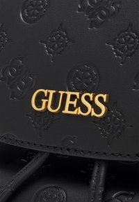 Černá kožená taška s texturou, zdobená embosovanými vzory a výrazným zlatým logem "GUESS". Detaily šití podtrhují design.