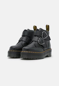 Bottines noires avec deux sangles ajustables, accessoires en forme de cœur, en cuir texturé, et une semelle épaisse à rainures avec des coutures jaunes.