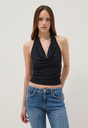 Jeune femme portant un haut noir sans manches à col halter et un jean bleu avec des clous décoratifs argentés à la ceinture et aux poches.