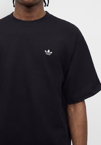 Άτομο που φοράει μαύρο T-shirt Adidas με μικρό λευκό λογότυπο trefoil στο αριστερό στήθος, με απλό ανοιχτό φόντο.