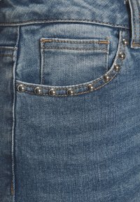 Zilas džinsi ar izbalinātu apdari, ar kabatu, kas rotāta ar sudraba kniedēm pa malu un kontrastējošu diegu.