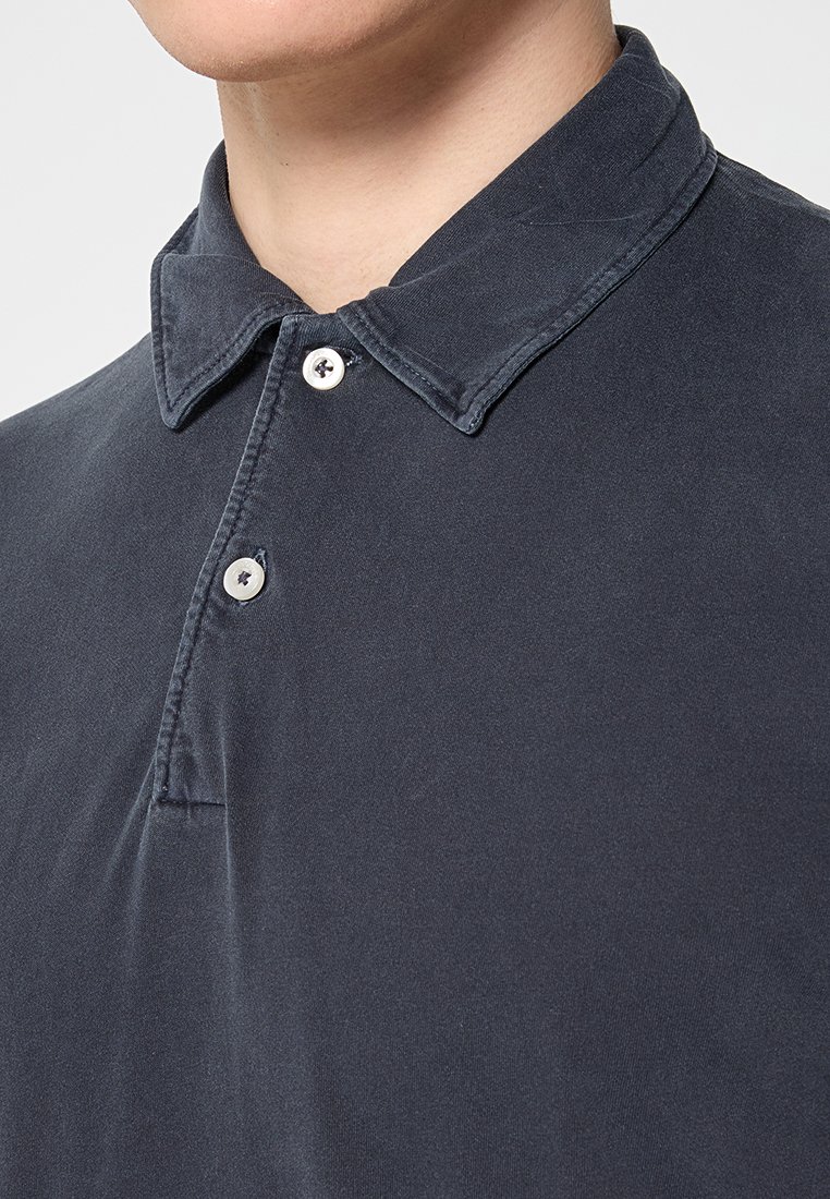 Mann trägt ein dunkelgraues Poloshirt mit zwei weißen Knöpfen, die am Kragen geschlossen sind, sichtbar vom Hals bis zur Brust.