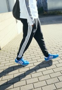 Sorte træningsbukser med hvide striber, prydet med et logo, sammen med blå sneakers med hvide detaljer, gående på en struktureret belægning.