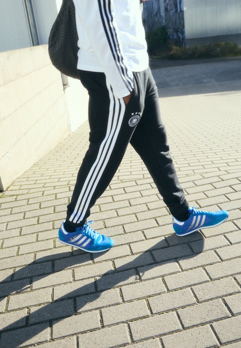 Sorte træningsbukser med hvide striber, prydet med et logo, sammen med blå sneakers med hvide detaljer, gående på en struktureret belægning.