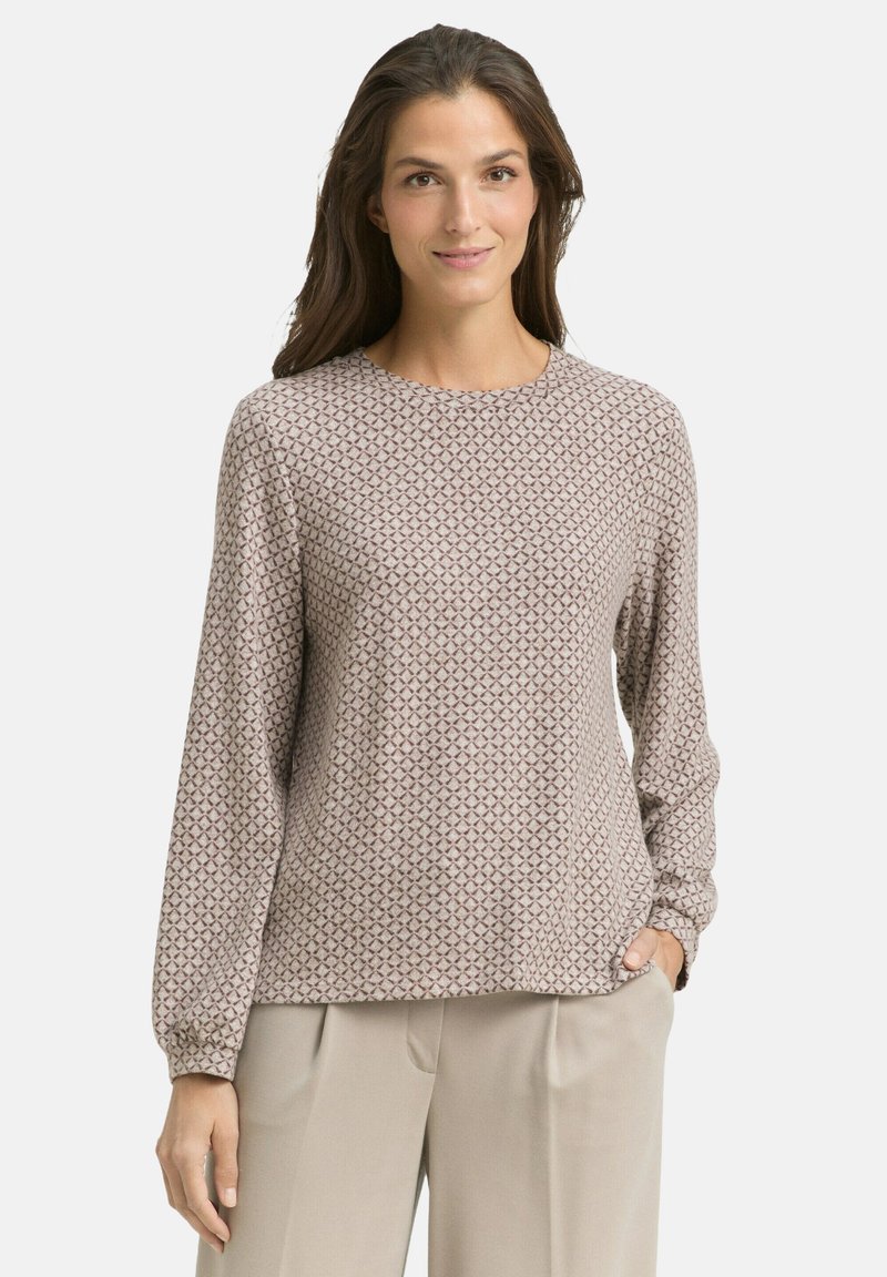 Vrouw in een lange mouwen beige blouse met patroontjes en ronde hals, draagt lichtbeige high-waisted broek en staat met één hand in haar zak.
