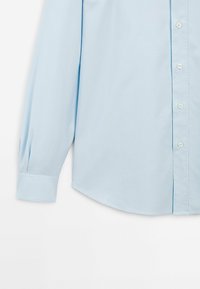 Camicia a maniche lunghe celeste, con bottoni frontali, orlo arrotondato, tessuto liscio e dettagli plissettati sulle maniche. Bottoni di colore a contrasto.