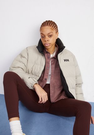 Dikke, tweekleurige lichtgrijze en zwarte jas met logo, gelaagd over een bruine shirt en witte top, gecombineerd met een paar bruine leggings op een blauwe ondergrond.