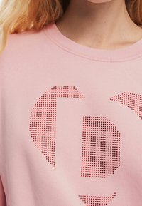 Rosa sweatshirt med rundad halsringning och ett mönster av röda prickar som formar bokstäver. Materialet verkar mjukt med en slät yta.
