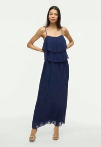 Moteris stovi su rankomis už nugaros, vilkinti tamsiai mėlyną klostuotą maksi suknelę su spaghetti petnešėlėmis ir aukso atvirais batais su kulnais.