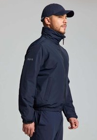 Giacca a vento blu navy con colletto alto, chiusura con zip, tasche laterali e polsini elasticizzati. Abbinata a pantaloni e cappellino blu navy coordinati.