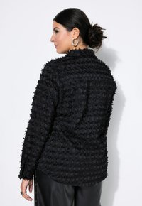 Camicia nera con texture caratterizzata da un design sfilacciato, con maniche lunghe e colletto, indossata sopra pantaloni neri eleganti. Tessuto morbido, vestibilità strutturata.